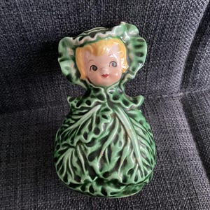 Puede incluir: Figura de cerámica vintage de una niña con un vestido y un gorro de hojas de col. La figura tiene el pelo rubio, mejillas rosadas y labios rojos. El vestido y el gorro son verdes con patrones de hojas detallados.