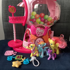 Op de afbeelding: Een roze My Little Pony kauwgomballenmachine speelgoedset met verschillende ponyfiguren en accessoires. De kauwgomballenmachine heeft een heldere bol gevuld met kleurrijke ballen. De set bevat een lollystandaard en diverse kleine speeltjes.