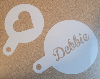 Könnte beinhalten: Zwei weiße Kunststoff-Schablonen zum Dekorieren von Kaffee oder anderen Getränken. Eine Schablone zeigt eine Herzform und die andere den Namen "Debbie" in kursiver Schrift.