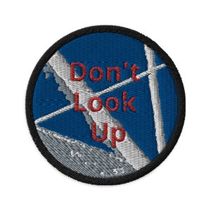 May include: Round embroidered patch featuring the text "Don't Look Up" in red. The patch has a blue background with a white and grey design, and a black border.