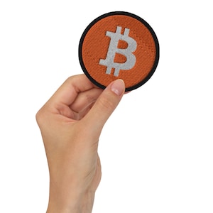 May include: Round orange embroidered patch featuring a white Bitcoin symbol, with a black border. The patch is held up by a hand against a white background. The Bitcoin symbol is a stylized B with two vertical lines.