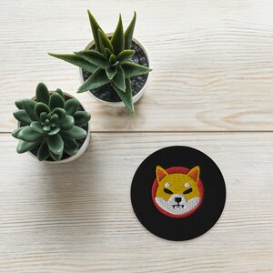 Könnte beinhalten: Runder, schwarz gestickter Aufnäher mit dem Shiba Inu Hundelogo in Gelb und Rot. Der Aufnäher liegt auf einer hellen Holzoberfläche neben zwei kleinen Topf-Sukkulenten.