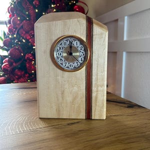 May include: A wooden clock with a light-colored wood body and a gold-colored clock face. A thin, dark red stripe runs vertically down the front. The clock face has black Roman numerals and hands. The clock is on a wooden surface.