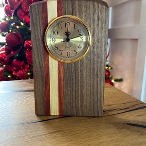 May include: A wooden desk clock with a gold-colored clock face. The clock has a rectangular shape with a dark brown wood grain. A vertical stripe of red, gold, and light wood accents the front. The clock face displays black numbers and hands.