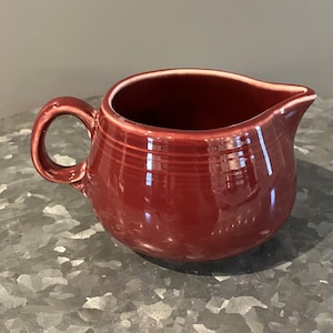 May include: A deep red ceramic creamer with a rounded body and a single handle. The creamer has a smooth, glossy finish.