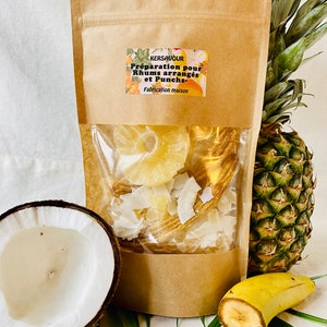 Peut inclure: Un sac en papier brun avec une étiquette indiquant "Kersavour Préparation pour Rhums arrangés et Punchs Fabrication maison". Le sac contient de l'ananas, de la banane et de la noix de coco séchés. Un ananas, une banane et une noix de coco sont visibles à l'extérieur du sac.