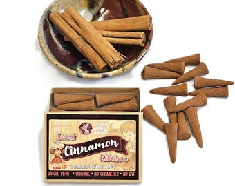 May include: A still-life image featuring cinnamon sticks in a decorative bowl and a box of handmade incense cones. The box is labeled "Sweet Cinnamon Kitchen" with a cartoon illustration. The cones are brown and cone-shaped.