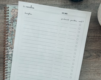 May include: A white notepad with the words "à vendre" and lined sections for descriptions, website, and pricing details. The notepad is on top of a spiral-bound world map notebook. A candle is in the background.