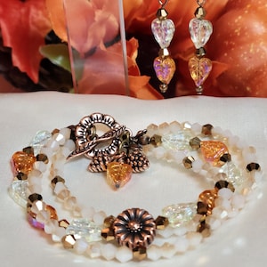 May include: A multi-strand bracelet and matching drop earrings set featuring white and amber-colored faceted crystal beads, copper-toned metal accents, a floral toggle clasp, and small dangling charms shaped like leaves and acorns.