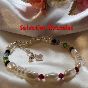 May include: A white pearl bracelet with a silver clasp and a small silver heart charm. The bracelet also features colourful glass beads and red crystal beads.