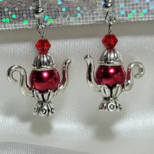 May include: A pair of earrings featuring miniature red teapot charms with silver-colored accents. Each charm has a red bead at the top and decorative detailing. The earrings hang from silver-colored hooks.