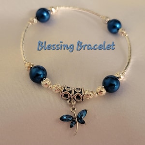May include: A silver bracelet with dark blue beads and a dragonfly charm. The bracelet has the words "Blessing Bracelet" in blue script. The dragonfly charm is dark blue with silver accents.