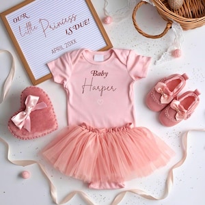 May include: A baby girl's announcement set featuring a pink onesie with "Baby Harper" printed on it, a pink tutu, and matching shoes. A heart-shaped pillow and a sign that reads "Our Little Princess is Due!" are also included.