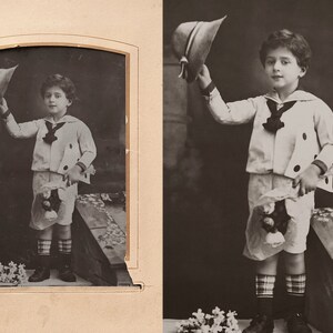 Pu&ograve; includere: Una fotografia in bianco e nero di un bambino piccolo che indossa un abito da marinaio e tiene un cappello. &Egrave; in piedi davanti a uno sfondo con fiori.