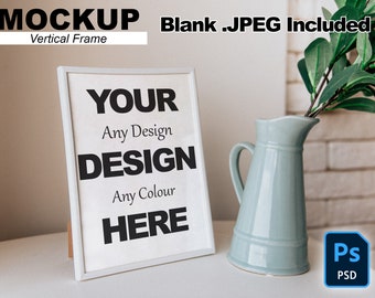 Op de afbeelding: Een witte verticale frame mockup met de tekst "YOUR DESIGN HERE" in zwarte tekst. De frame staat op een witte tafel met een lichtblauwe keramische kan en een groene plant op de achtergrond.