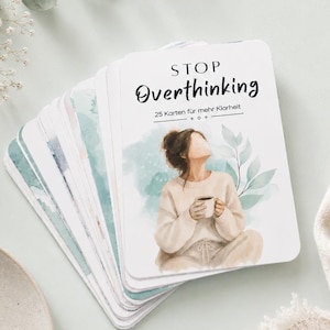 Può includere: Un mazzo di carte rettangolari con angoli arrotondati, recante il testo STOP Overthinking e 25 Karten für mehr Klarheit. La carta superiore mostra un'illustrazione ad acquerello di una persona con un maglione beige che tiene una tazza, con foglie verdi.