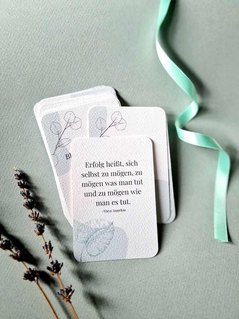 May include: A stack of white cards with rounded corners, featuring a light blue botanical design and text in German. The cards are arranged on a light green surface, with a sprig of lavender and a green ribbon in the background.