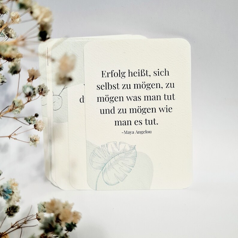 May include: A stack of off-white cards with rounded corners, featuring a quote in German by Maya Angelou and a light blue leaf illustration. The cards are surrounded by delicate, dried flowers.