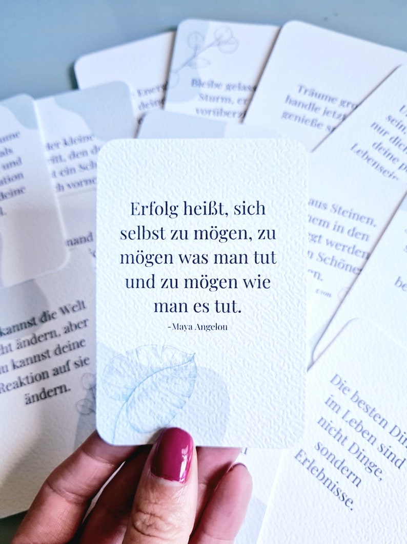 May include: A deck of inspirational cards with a light blue floral design. One card features a quote by Maya Angelou in black text. Other cards with text are visible in the background.