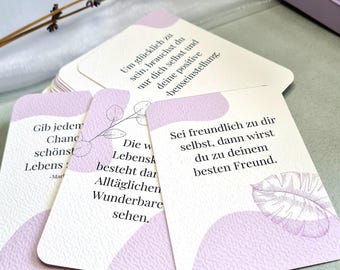 May include: A set of purple affirmation cards with inspirational quotes in German. The cards feature a white background with a light purple border and a light purple abstract shape. The text on the cards is in black.