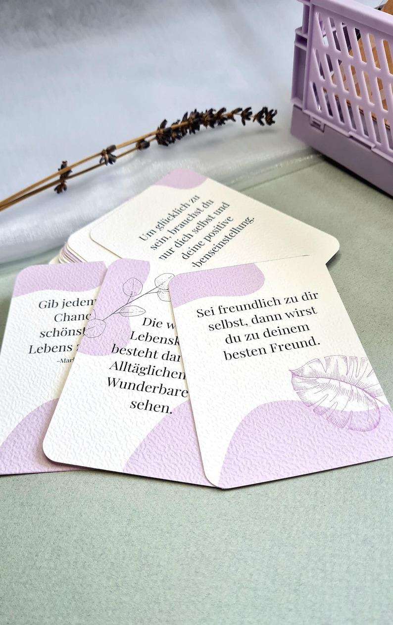 May include: A stack of inspirational cards with rounded corners, featuring soft lavender accents and handwritten text. The cards have quotes and phrases. A lavender storage bin and dried flower sprig are in the background.