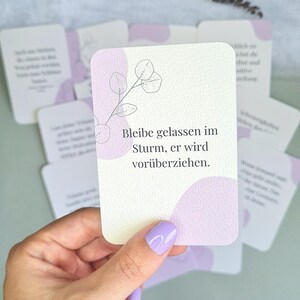 May include: A deck of affirmation cards with a light purple and white design. The cards have a floral illustration and the German text "Bleibe gelassen im Sturm, er wird vor&uuml;berziehen." The cards are held in a hand with purple nail polish.