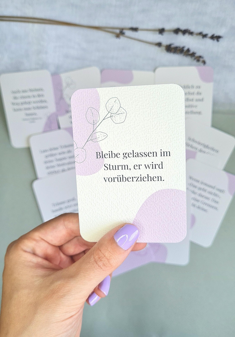 May include: A deck of affirmation cards with a light purple and white design. The cards have a floral illustration and the German text "Bleibe gelassen im Sturm, er wird vor&uuml;berziehen." The cards are held in a hand with purple nail polish.