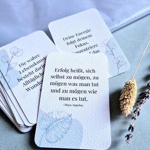 May include: A stack of white cards with rounded corners, featuring inspirational quotes and delicate botanical illustrations in blue. The cards have a textured surface and are arranged on a light blue background, with a sprig of lavender and dried flower.