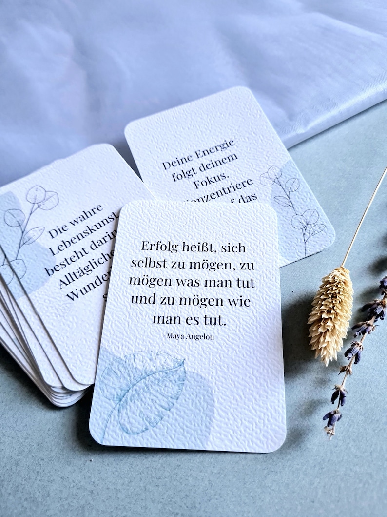 May include: A stack of white cards with rounded corners, featuring inspirational quotes and delicate botanical illustrations in blue. The cards have a textured surface and are arranged on a light blue background, with a sprig of lavender and dried flower.