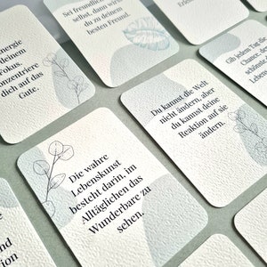 May include: A collection of white cards with inspirational quotes in German. Each card features a textured surface, soft blue watercolor-style shapes, and delicate botanical illustrations. The cards are arranged on a light green surface.