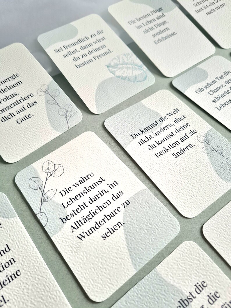 May include: A collection of white cards with inspirational quotes in German. Each card features a textured surface, soft blue watercolor-style shapes, and delicate botanical illustrations. The cards are arranged on a light green surface.