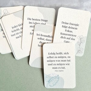 May include: A collection of cream-colored affirmation cards with rounded corners, featuring inspirational quotes in German. Each card has a subtle watercolor design in shades of blue and gray. The cards are arranged on a light green surface.