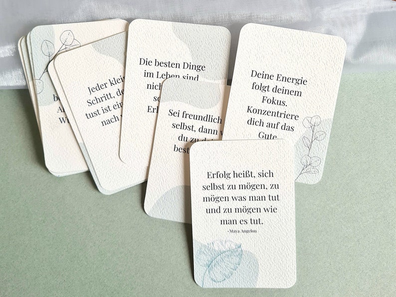 May include: A collection of cream-colored affirmation cards with rounded corners, featuring inspirational quotes in German. Each card has a subtle watercolor design in shades of blue and gray. The cards are arranged on a light green surface.