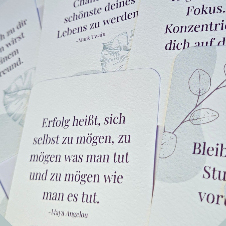 May include: A collection of white cards with inspirational quotes in German. The cards feature botanical illustrations in a soft green hue. Quotes include phrases by Mark Twain and Maya Angelou. The cards are arranged in a slightly overlapping manner.
