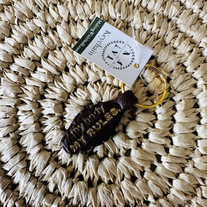 May include: A dark brown leather keychain with the words "MY BODY MY RULES" in gold lettering. The keychain is attached to a gold-colored ring and a white tag that reads "Keychain" and "MakingWithMollie.com". The background is a woven straw mat.