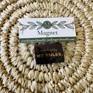 May include: A dark brown magnet with gold lettering reading "my body my rules" is displayed in a clear plastic sleeve. The magnet is rectangular. A white card with the word "Magnet" and the maker's social media handle is above the magnet.