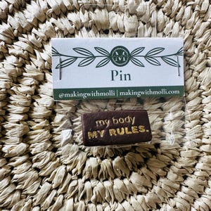 May include: A rectangular brown pin with the text "my body MY RULES" in gold lettering. The pin is packaged on a white card with the word "Pin" and a logo. The background is a woven, beige material.