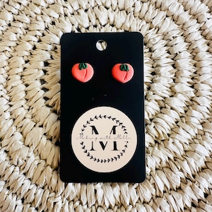 Pode incluir: Brincos de botão feitos à mão em forma de tomate vermelho com detalhes de folhas verdes, apresentados em um cartão preto. O cartão tem um logotipo circular com o texto "Making with Melle".