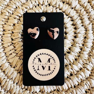 May include: A pair of heart-shaped stud earrings on a black card. The earrings are pink with black marbling. The card has a white sticker with the text "Making with Mille" in a circular design.