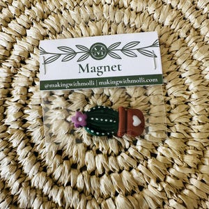May include: A cactus-shaped magnet in a clear plastic sleeve. The magnet is green with white dots, a purple flower, and a brown pot with a white heart. The packaging reads "Magnet" and "@makingwithmolli | makingwithmolli.com".