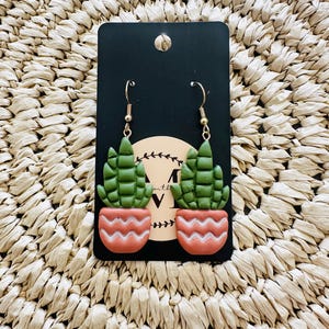 May include: A pair of handmade earrings featuring a green cactus design in a coral-colored pot. The earrings are attached to gold-colored hooks and displayed on a black card. The background is a woven, beige material.