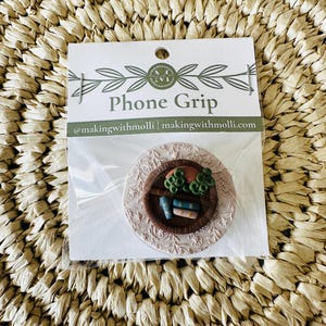Pode incluir: Um suporte para telemóvel com um diorama em miniatura de livros e vegetação. O suporte para telemóvel redondo tem uma base castanha com um design floral rosa claro. A embalagem diz "Phone Grip" e "@makingwithmolli | makingwithmolli.com".