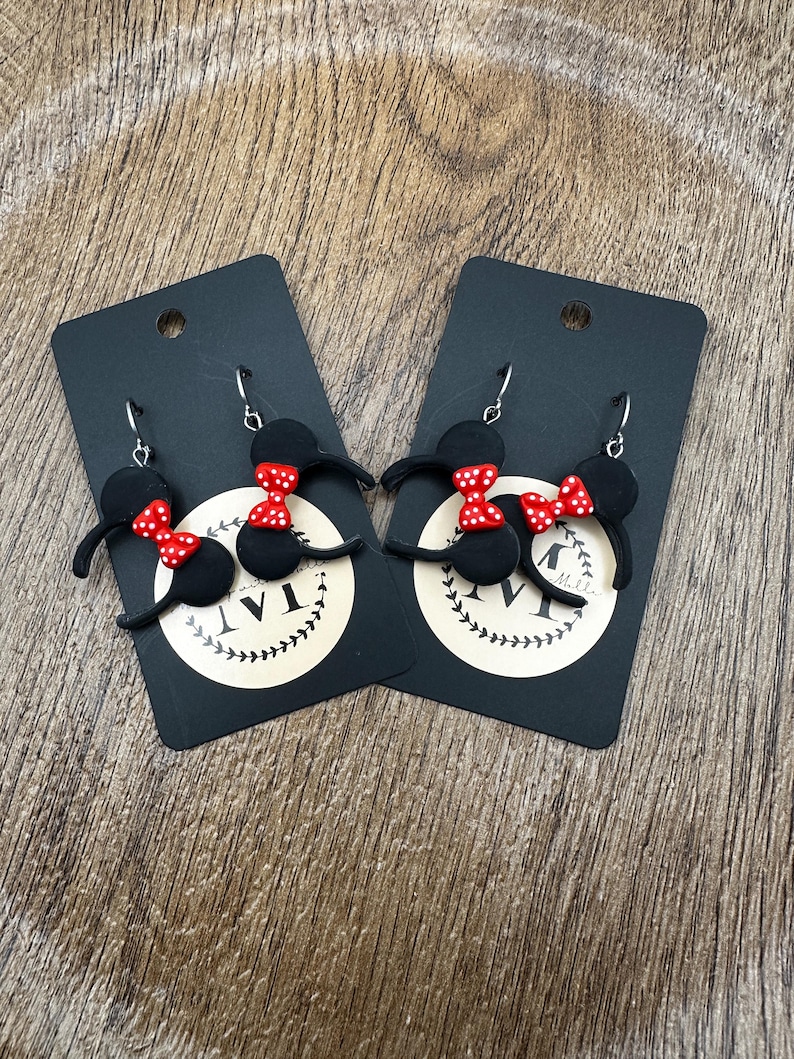 May include: Two pairs of black polymer clay earrings shaped like mouse ears with red polka dot bows. The earrings are on black cards with a white logo and silver earring hooks. The background is a wooden surface.