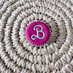 May include: A circular, hot pink phone grip featuring a scalloped edge and a prominent, white, stylized letter B centered on the surface. The accessory has a smooth, matte finish and a clear plastic base visible beneath the circular top piece.
