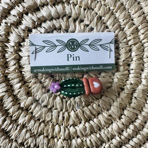 Puede incluir: Un pin hecho a mano con un cactus verde con puntos blancos, una flor morada y una maceta roja con un corazón blanco. El pin se muestra en una tarjeta que dice "Pin" y el identificador de redes sociales del creador.