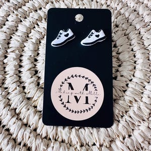 May include: A pair of white and black trainer-shaped stud earrings attached to a black rectangular display card. The card features a circular logo with a wreath design and the text Making with Molle MVI in the centre, with a hole at the top.
