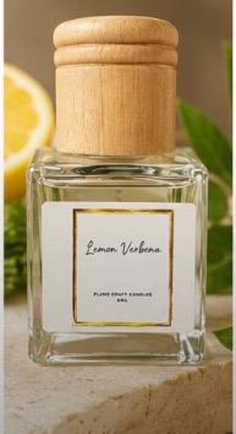 May include: A clear glass bottle with a light wood cap, containing a liquid. The white label features a gold border and the text Lemon Verbena, FLAME CRAFT CANDLES, 5ML. The bottle has a square base and a cylindrical neck with a wooden stopper.