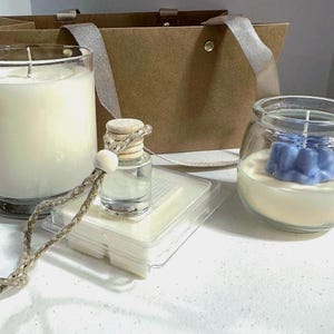 May include: A selection of scented candles and wax melts. A large white candle in a glass jar, a small blue flower-shaped candle, a small glass bottle, and a stack of wax melts are displayed with a brown paper gift bag.