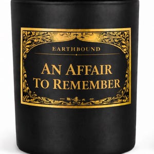 Op de afbeelding: Zwarte kaars met een houten deksel. De kaars heeft een gouden label met de woorden "EARTHBOUND" en "AN AFFAIR TO REMEMBER" in gouden letters. De kaars is cilindrisch.