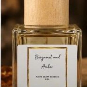 May include: A square glass bottle filled with a light amber liquid, topped with a cylindrical light wood cap. The white label features a gold border and the text Bergamot and Amber, Flame Craft Candles, 60ml.